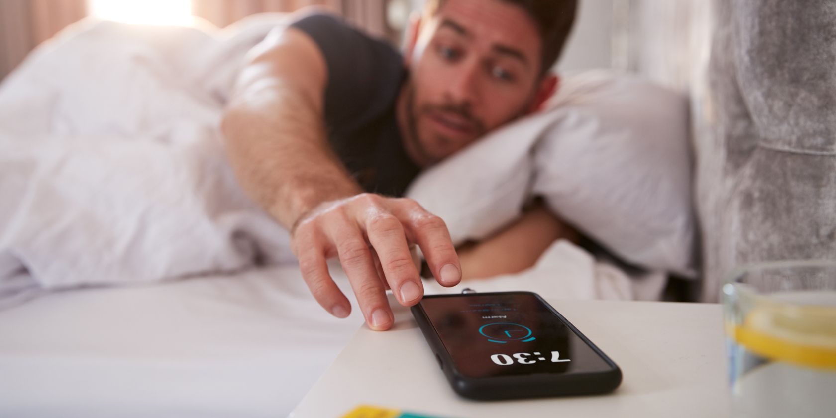 man in bed reaching for phone displaying alarm