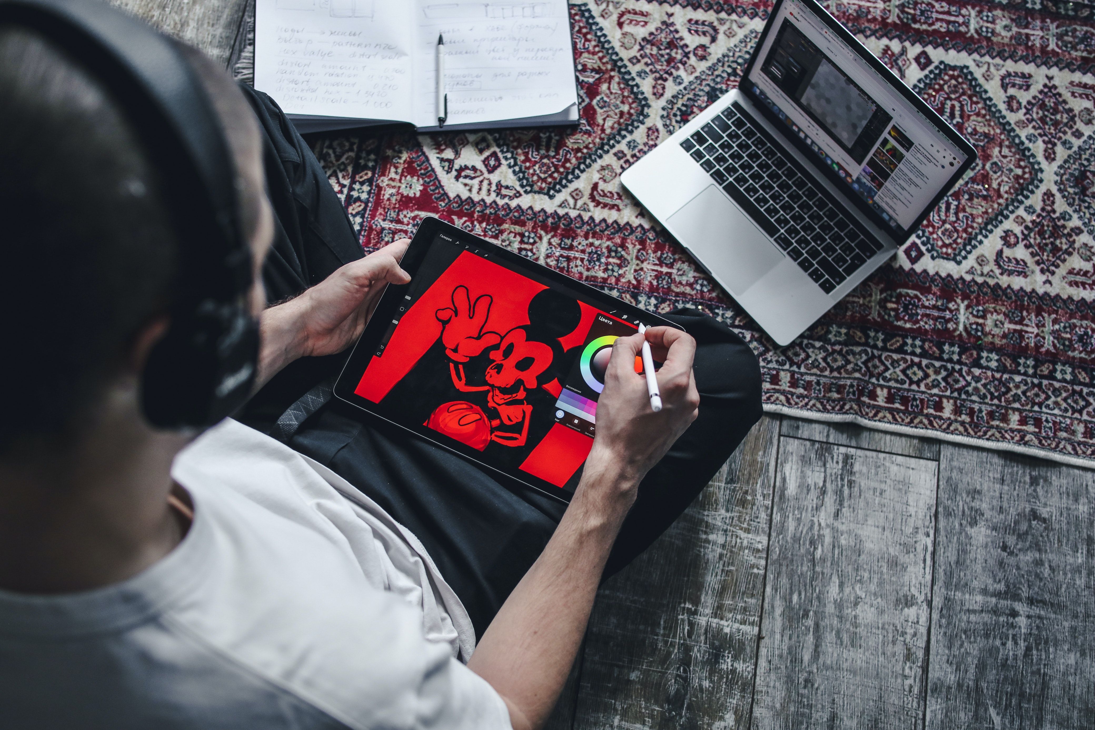 Man sitting on the floor using technology to sketch cartoons on an iPad while watching a laptop.