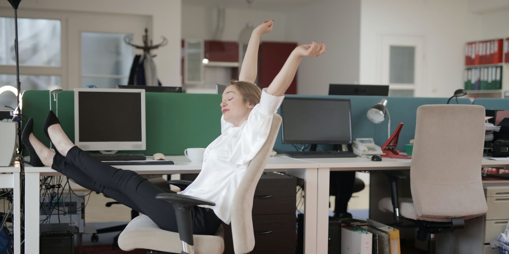 Ecstatic female employee in a tech office with her hands in the air and legs resting on the work table surrounded by desktop workstations.