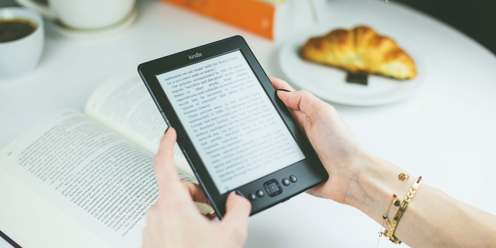 Photo of a Kindle being read by an unseen person with a book and a pastry on the table below