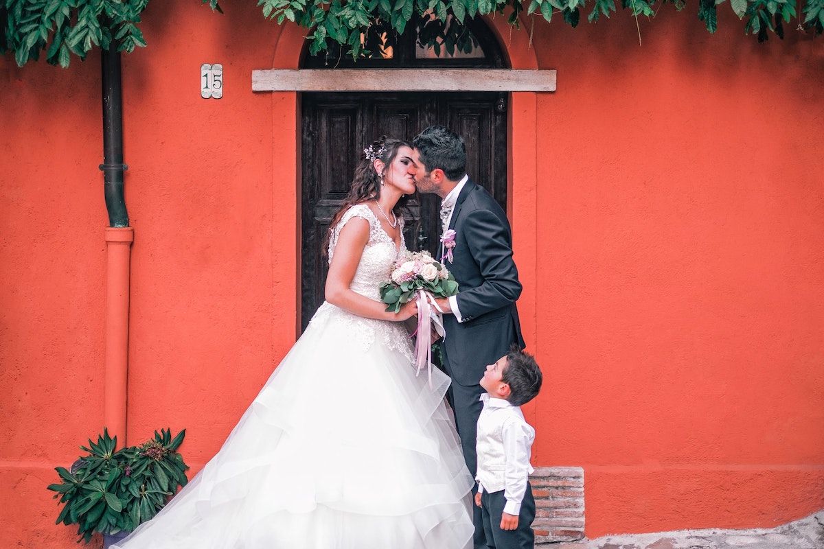 The bride and groom are kissing in front of a bright red wall, and a child is looking up.