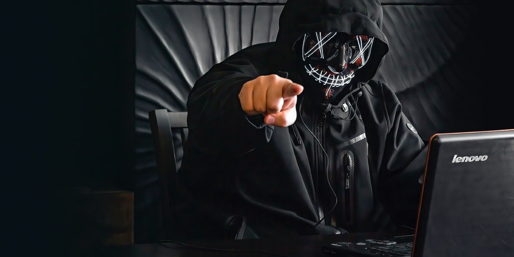 hacker in black hoodie and mask in front of a laptop pointing at the camera