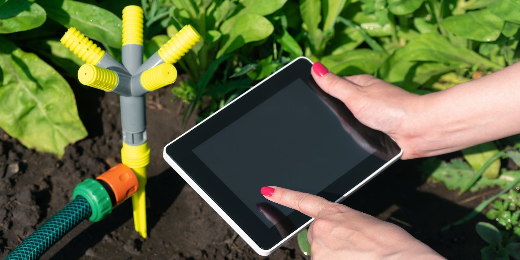 Two hands holding a tablet device next to a garden sprinkler system