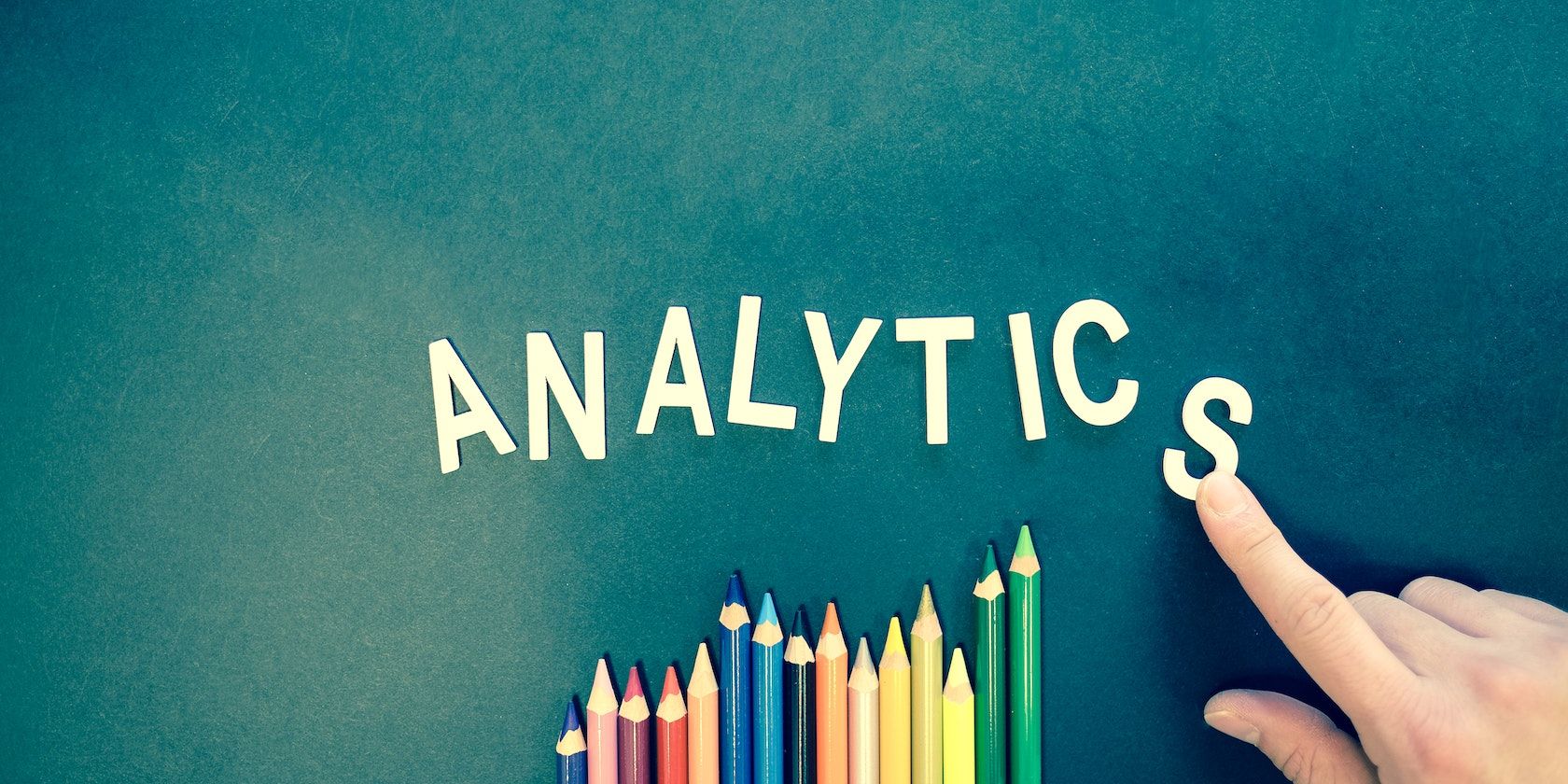 A finger arranges letters to spell the word 'Analytics' above a row of coloured pencils