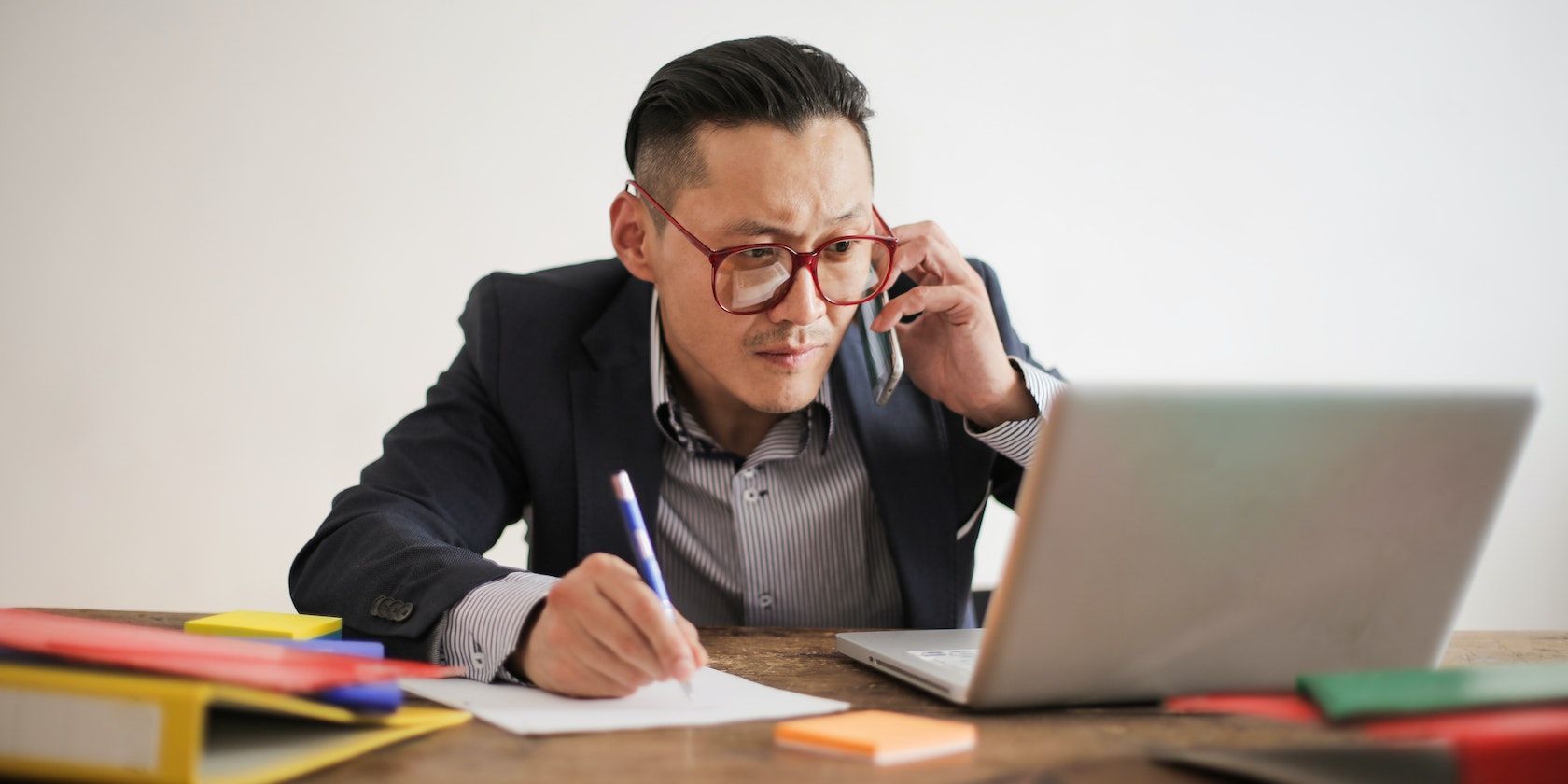 A man in a black suit, writing on a paper and staring at a laptop screen simultaneously.
