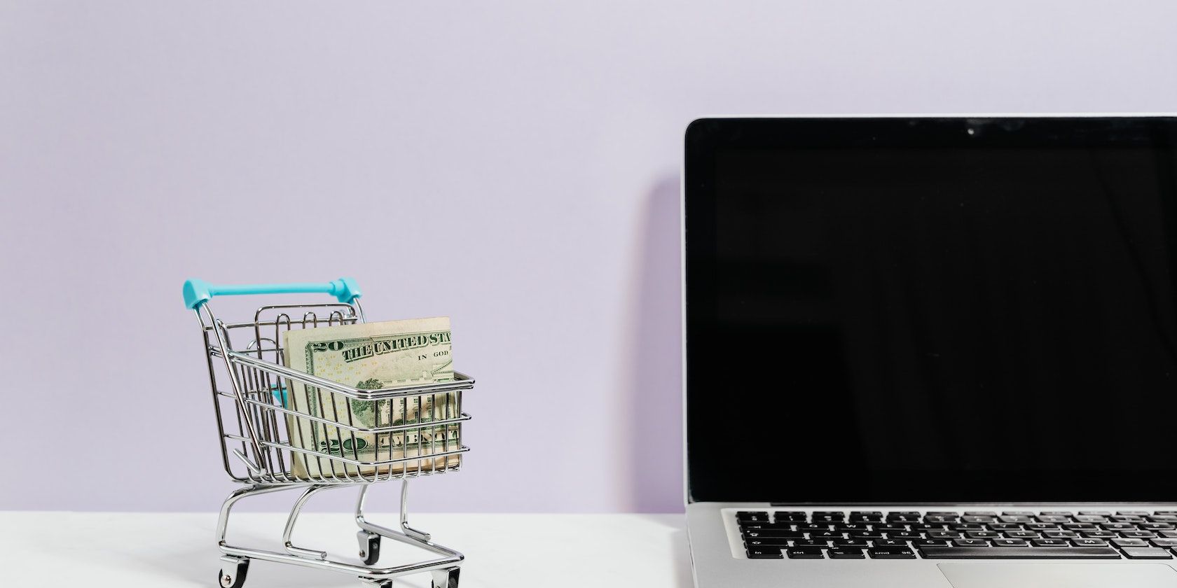 Macbook on White Table Beside a Miniature Shopping Cart With Money 