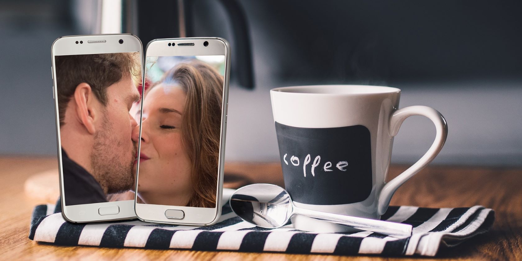 A man and woman's face on two smartphones kissing with a coffee cup on the table