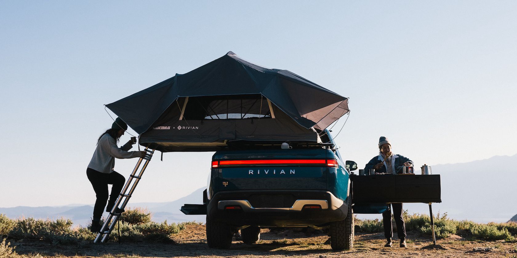 A blue Rivian R1T is set up for outdoor camping with a roof mounted tent