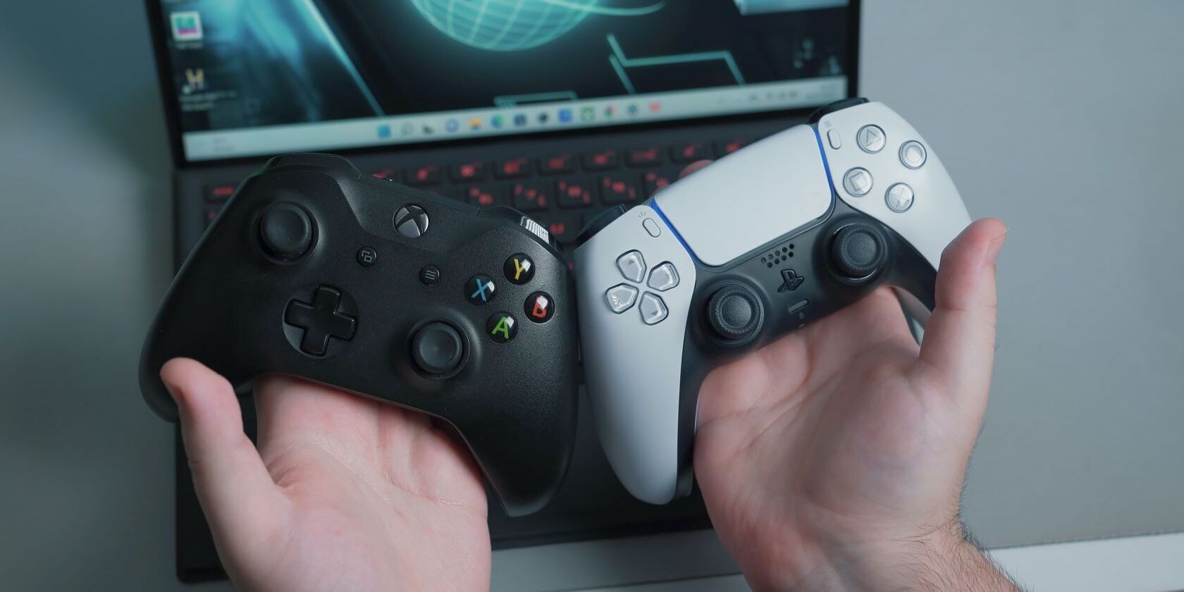 A photo of a man holding up a black Xbox One Controller and a standard PlayStation 5 controller in front of a laptop