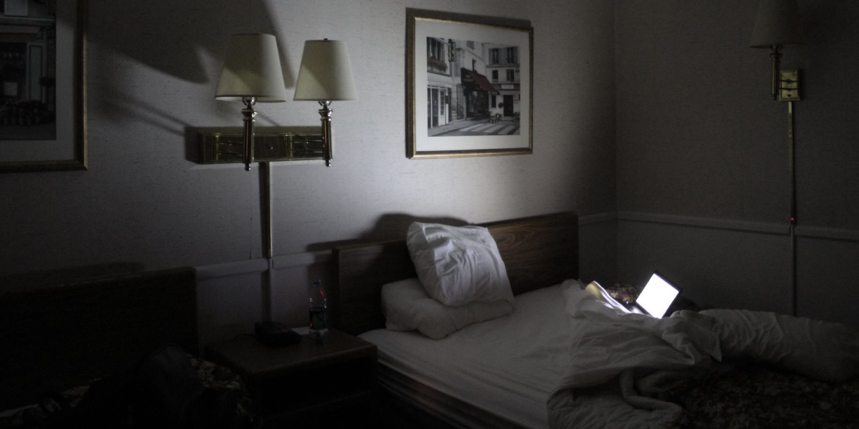 An open laptop on a bed illuminates an otherwise dark hotel room.