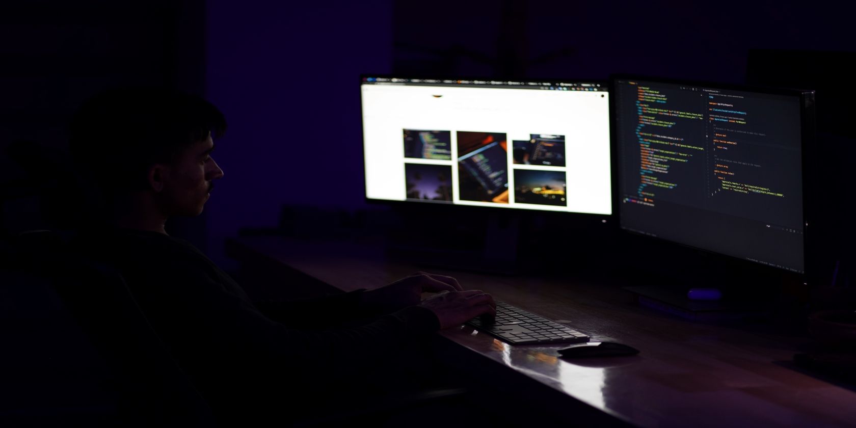Somebody sitting at a desk, in a very dark room, in front of two screens, one showing a web page, the other showing code in a text editor.