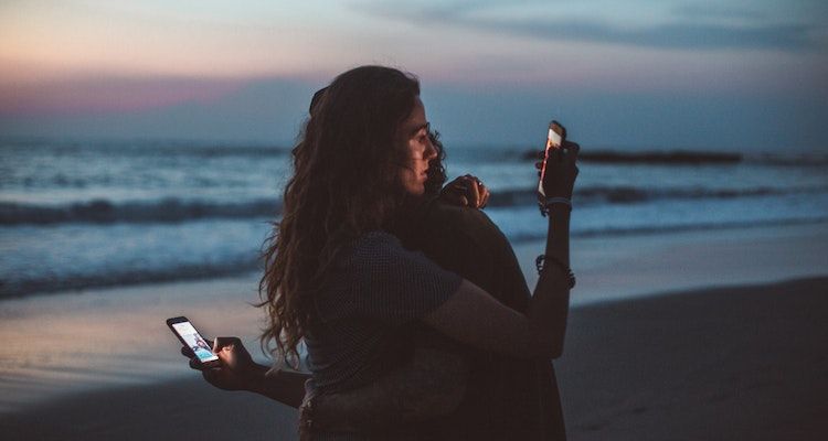 زن و شوهری در ساحل در آغوش گرفته و به تلفن هایشان نگاه می کنند