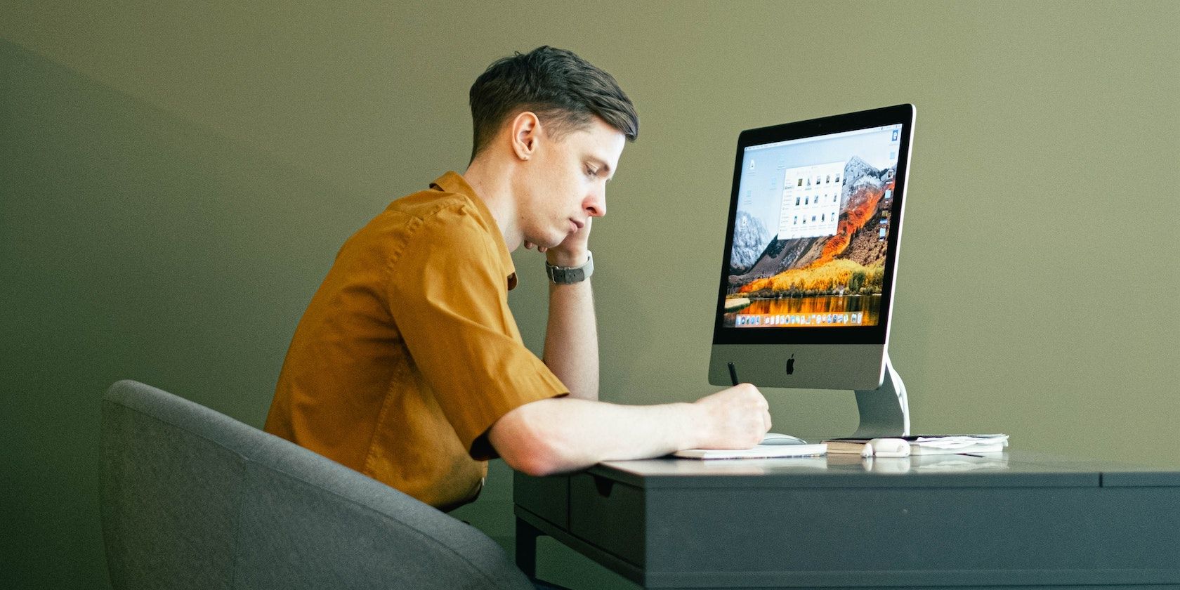 Man writing in a notebook while sitting in front of an iMac