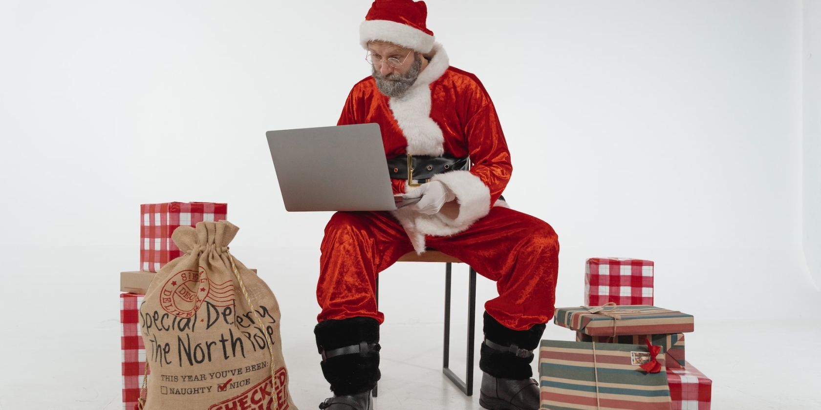 Père Noël assis sur une chaise avec un ordinateur portable entouré de cadeaux