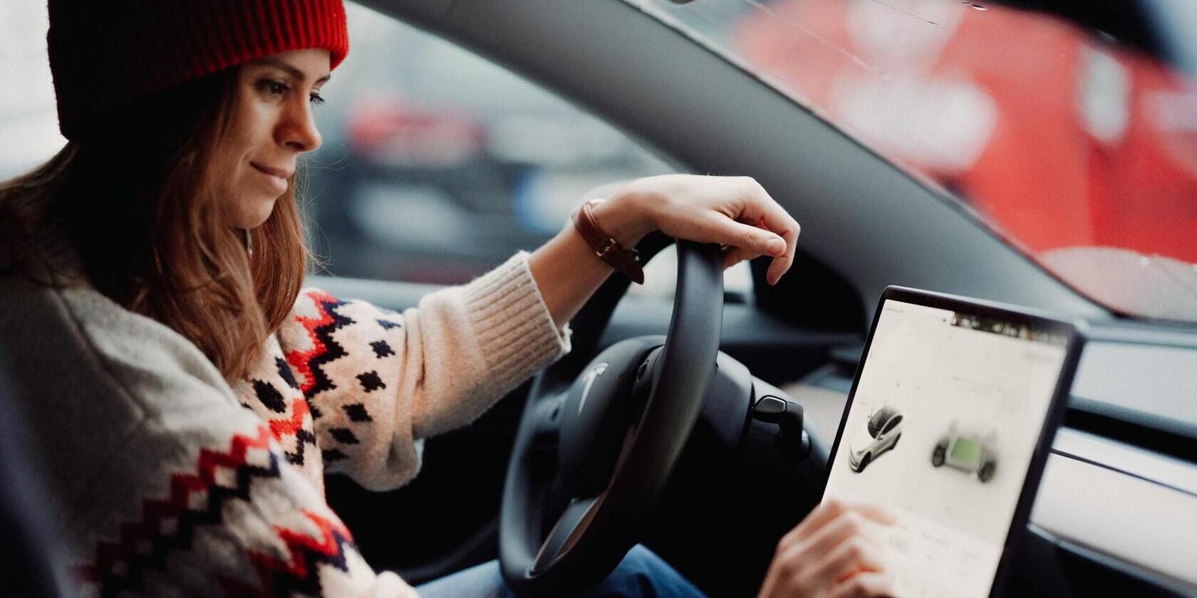 A Tesla driver wearing a sweater and hat uses their touchscreen. 