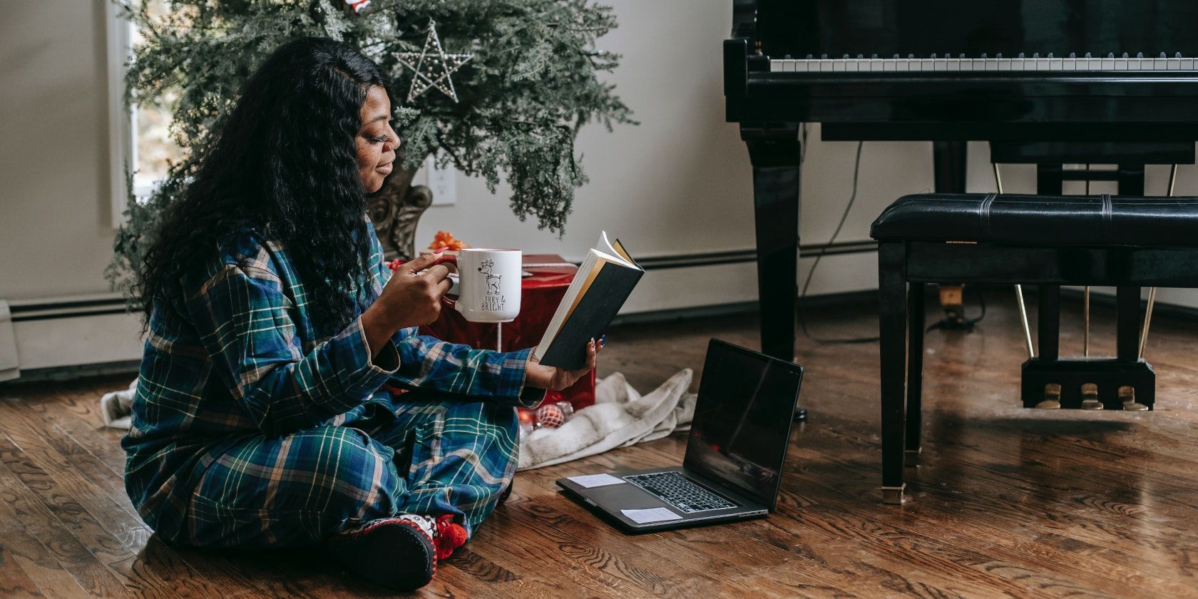 Mujer leyendo un libro junto a un árbol de Navidad y una computadora portátil