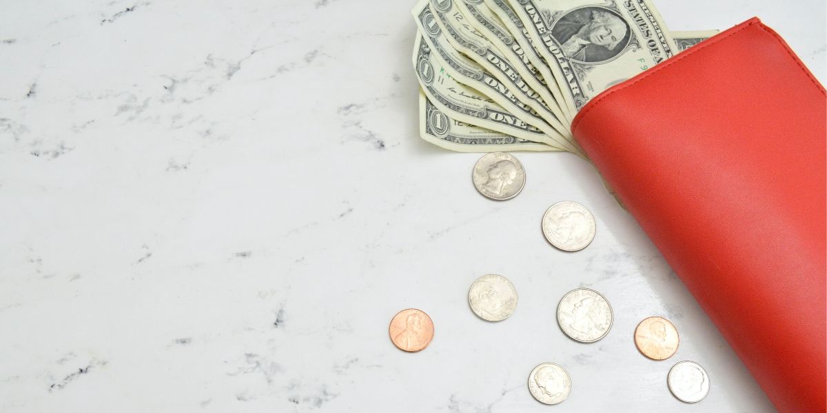 Red purse containing some dollars and coins on a marble table.