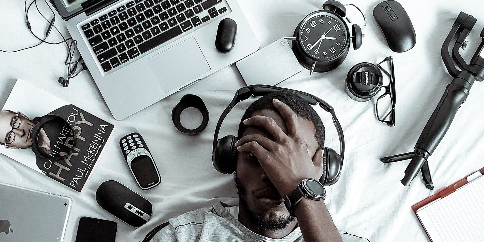 Man in black headphones covering his eyes surrounded by technology devices