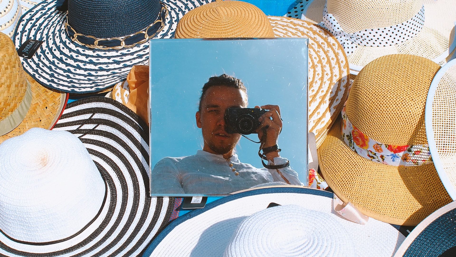 Man taking a self portrait using a mirror that is surrounded by summer hats.