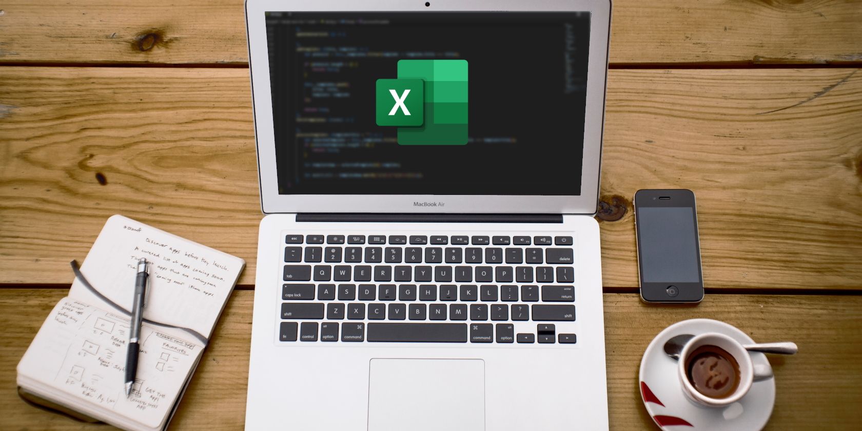 A Cup of Coffee, Phone, Notebook, and Computer on a table. The computer has code in the background of the screen and the Excel logo in the foreground. 