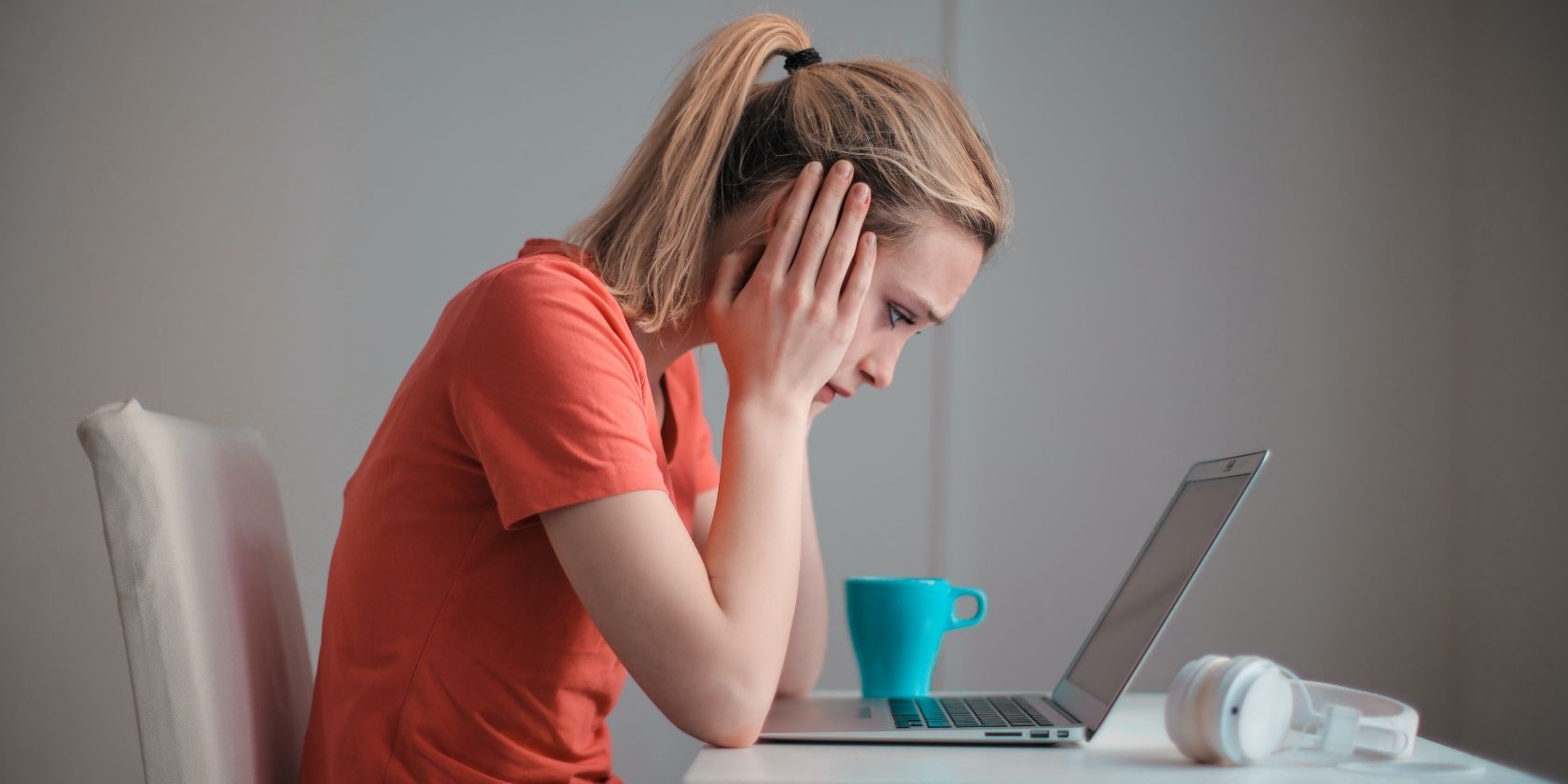 Blond woman with ponytail and orange shirt looking at her laptop with both hands on the sides of her face looking disappointed