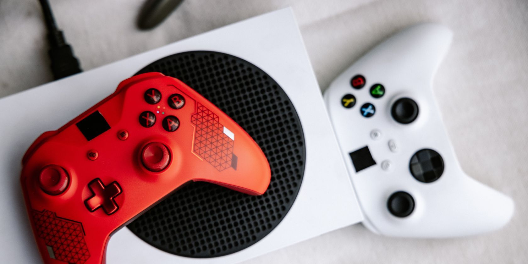 An Xbox One Series S with a red controller on top of it and a white controller nearby