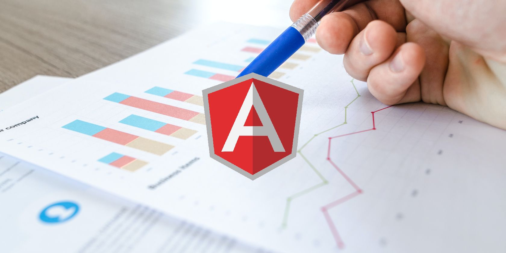 The Angular logo superimposed over a hand holding a pen above some paper containing charts and graphs