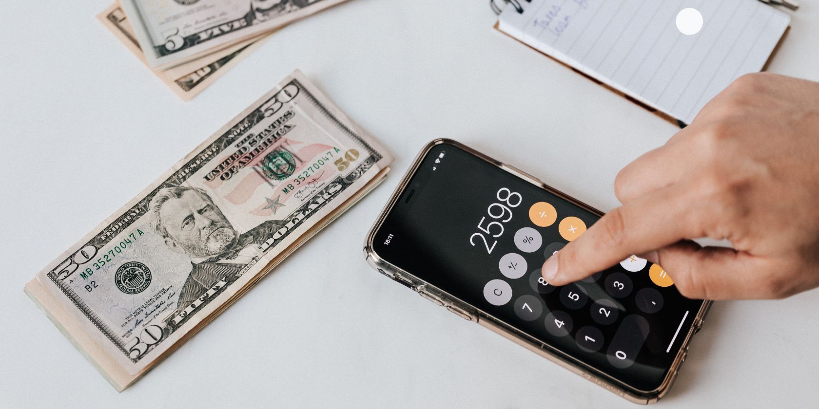 person using the calculator app on a smartphone placed on a table beside dollar bills and a notebook