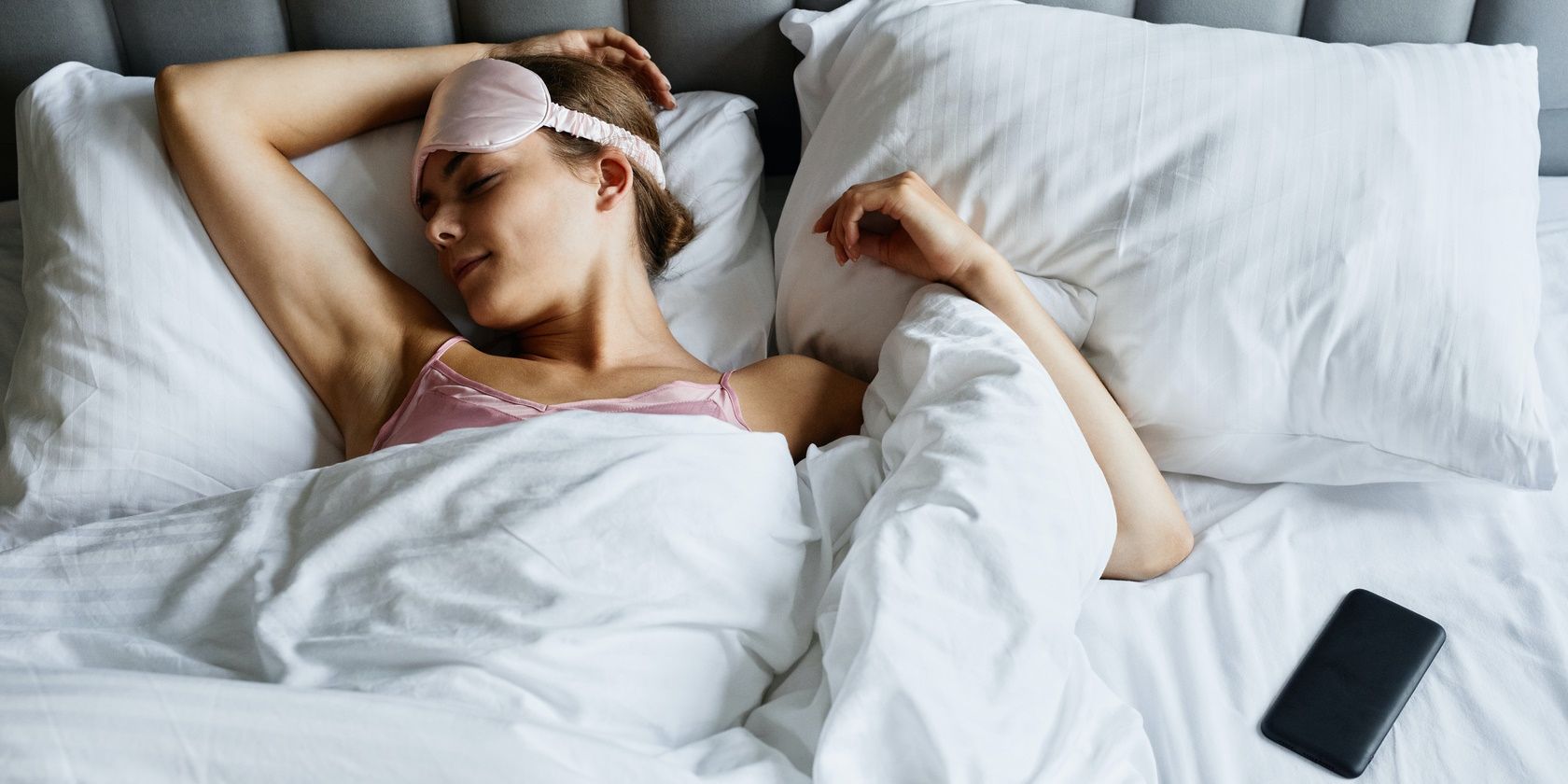 woman smiling sleeping wearing eyemask and lying next to black smartphone