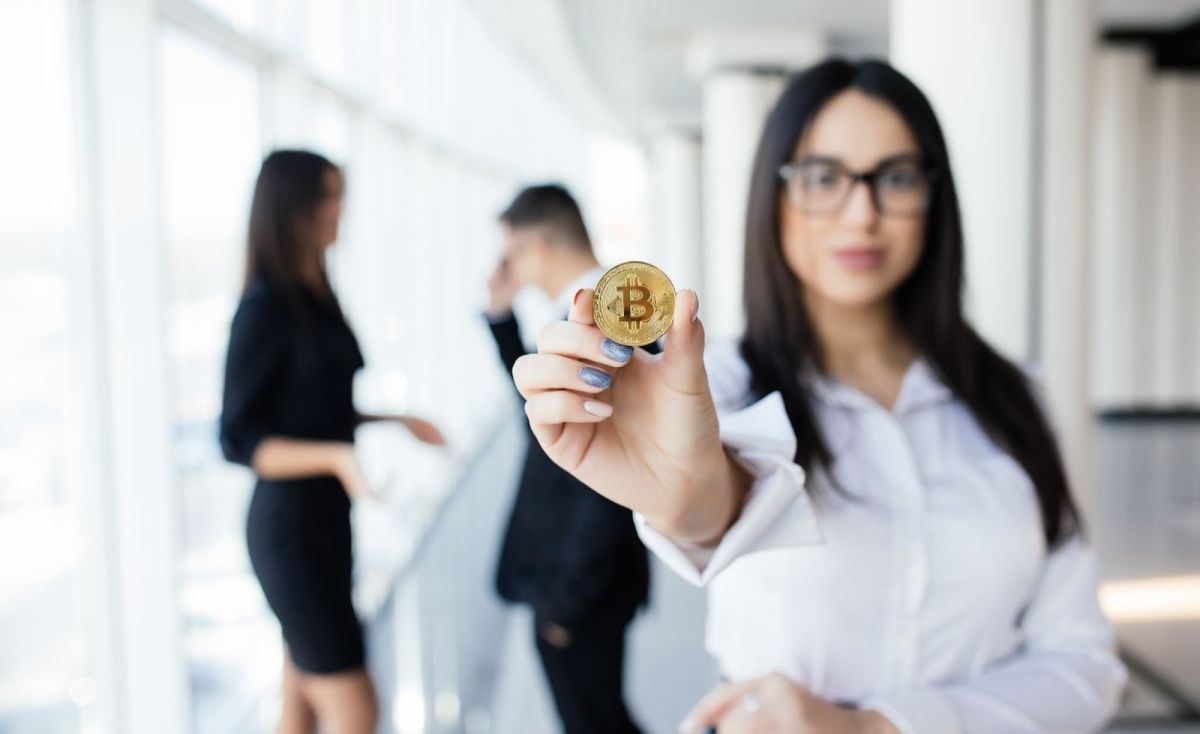 A businesswoman holding a gold BTC and two people doing a discussion in the background .