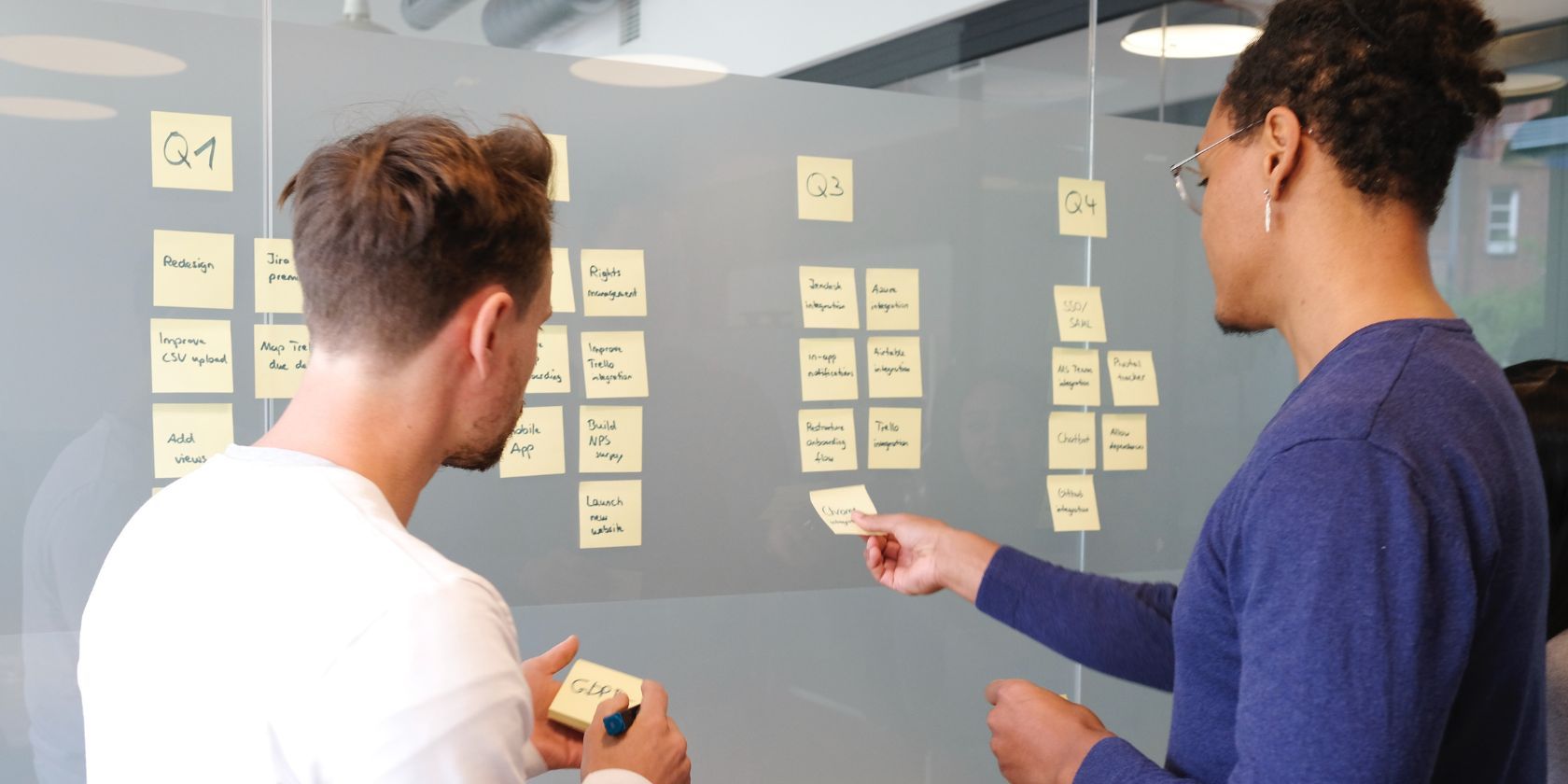 Two male co-workers organizing a to-do list on a glass wall with sticky notes