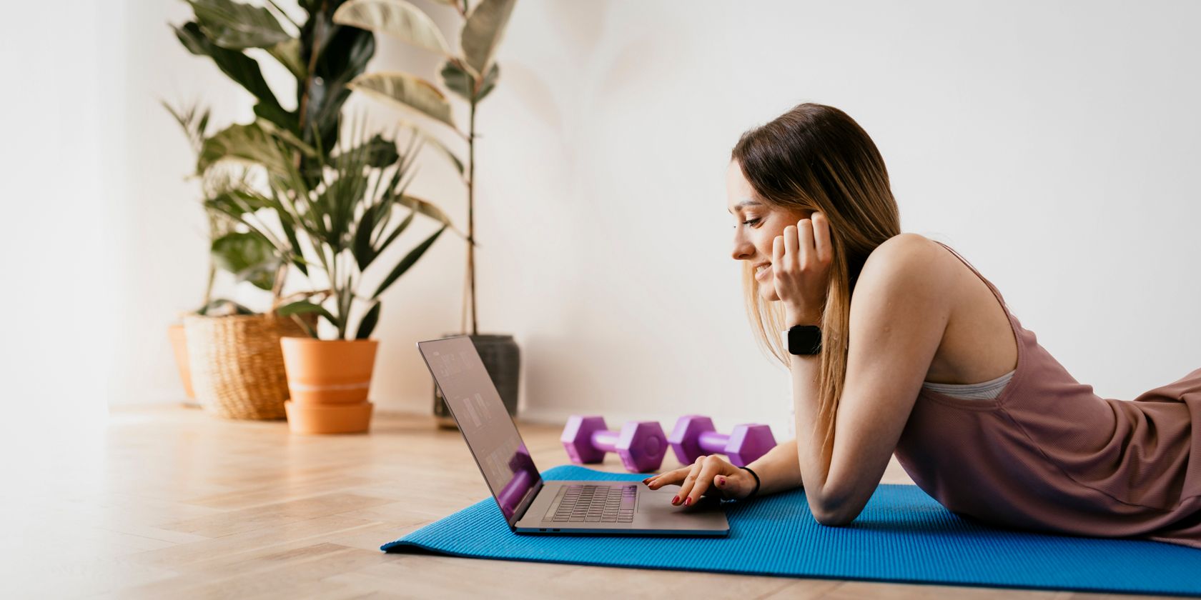 A woman lying on an exercise mat with weights choosing a workout on a laptop