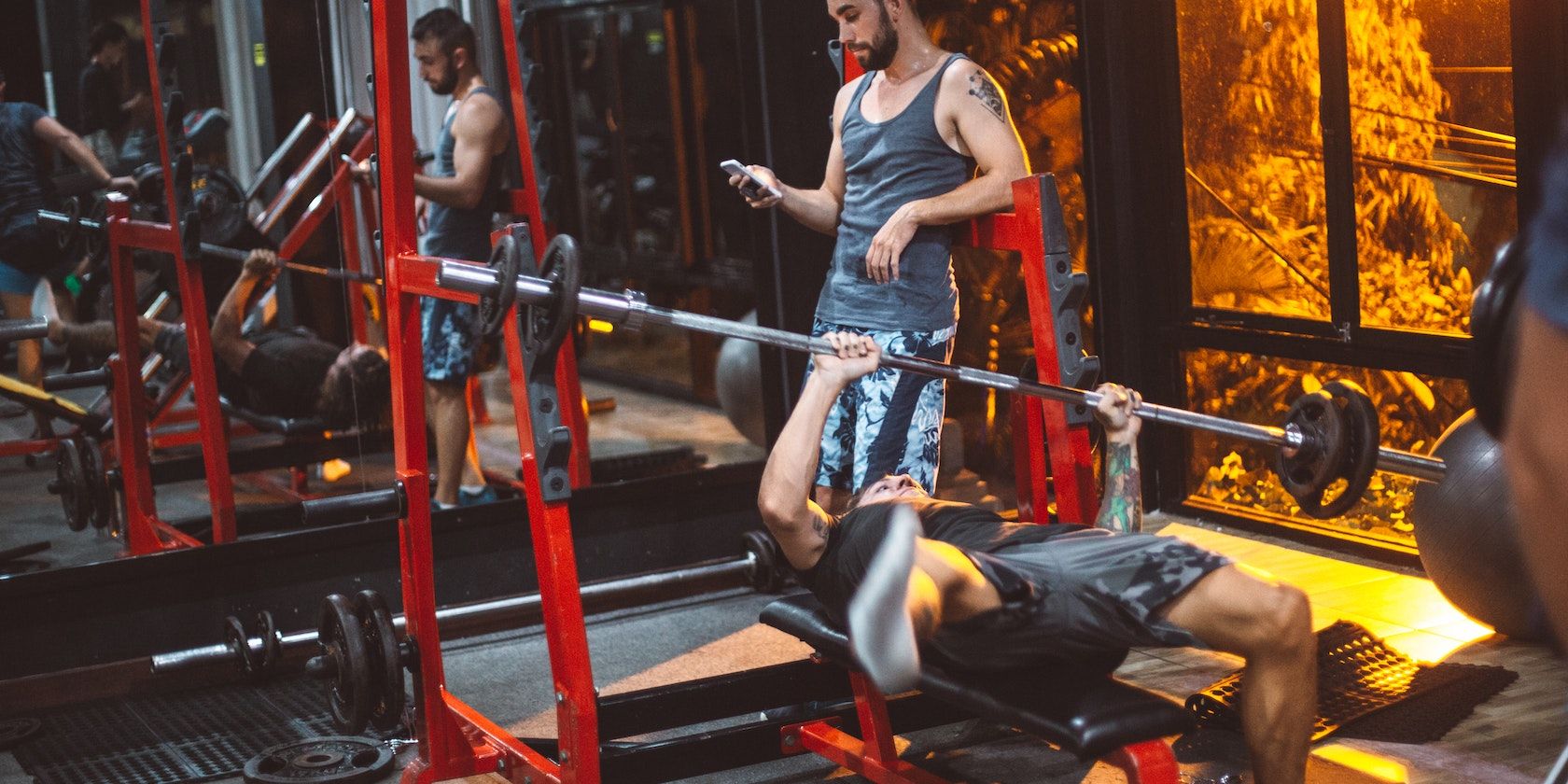 Man struggling with barbell bench press while spotter uses phone