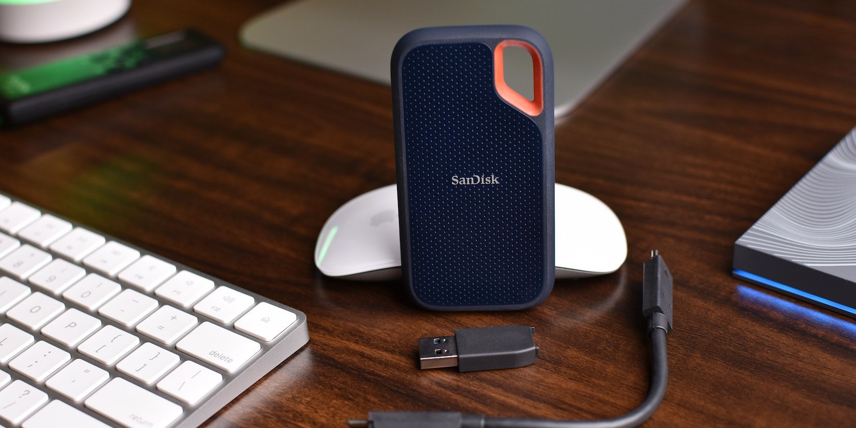 Sandisk SSD beside a USB drive, a cable, an Apple mouse, and a Magic Keyboard