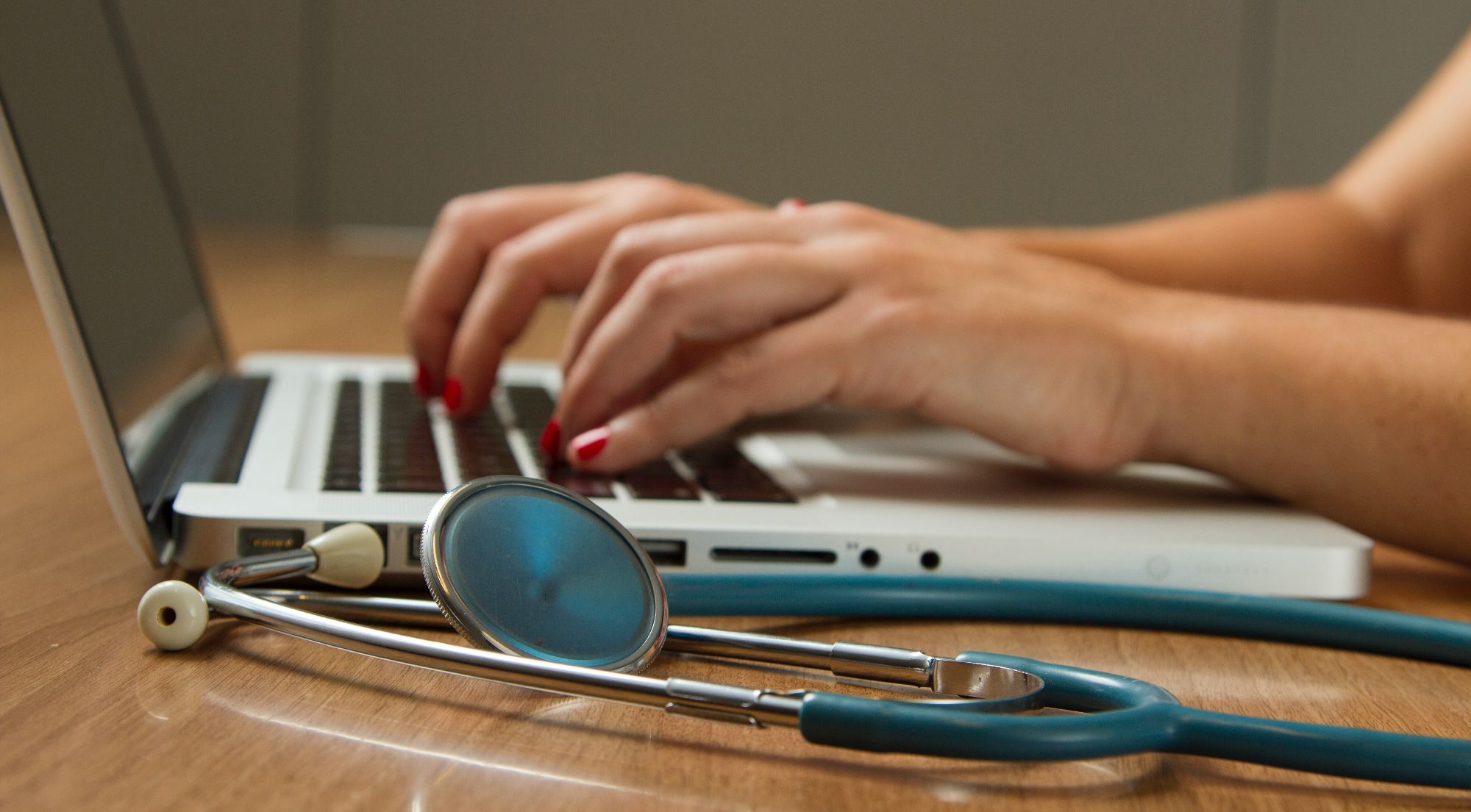 person typing on laptop next to stethoscope on desk