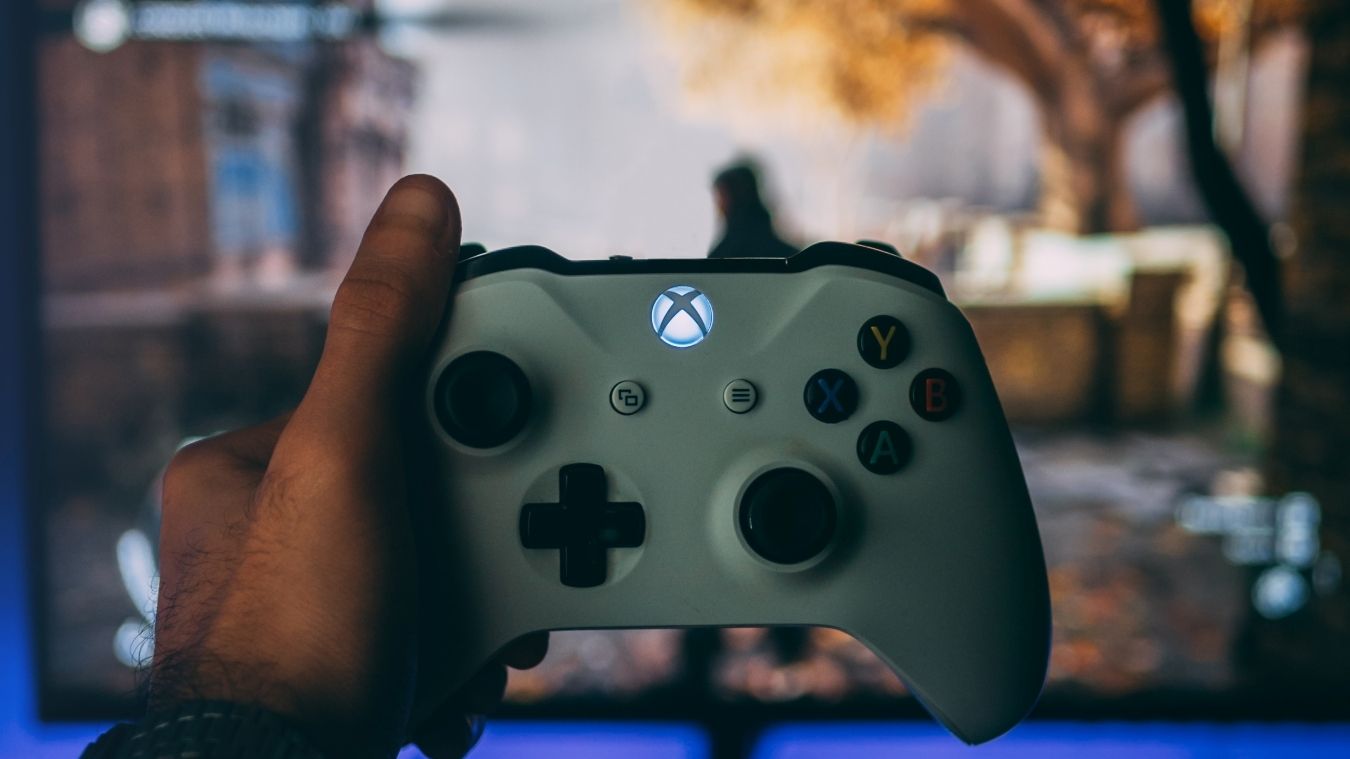 A photograph of a white Xbox Wireless Controller held in front of an out of focus monitor  