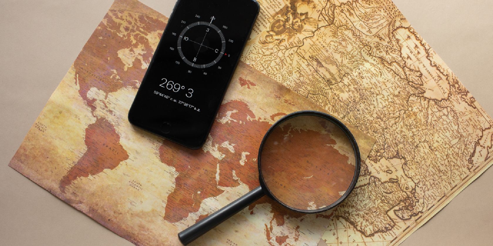 A magnifying glass and a phone display a compass app on top of two old-fashioned maps.