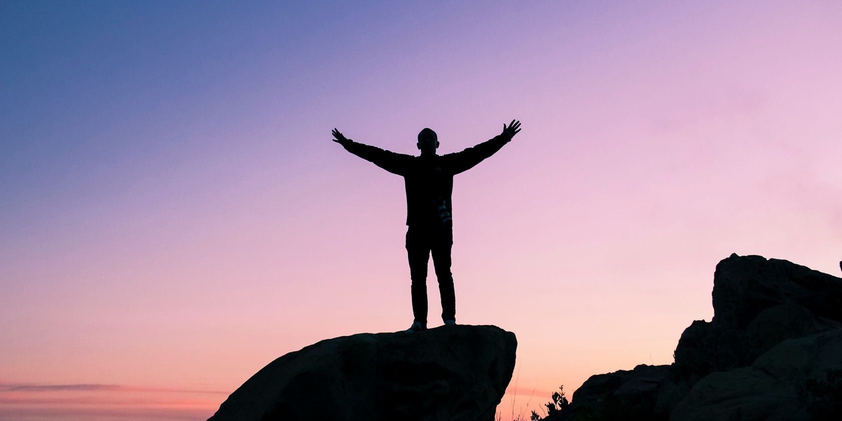 A man stands with arms outstretched at the top of a mountain at sunset