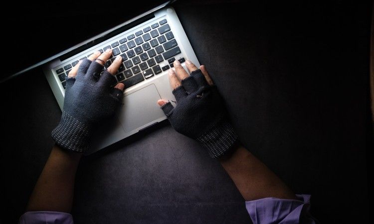 A man wearing dark, fingerless gloves trying to type of a laptop down a dark road.
