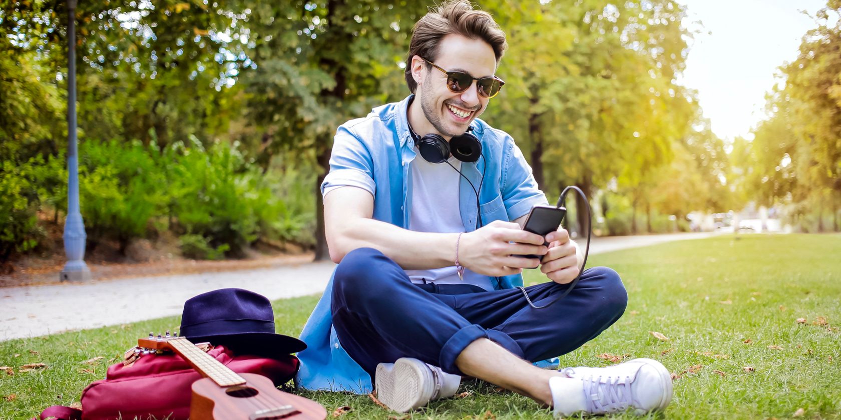 A happy man sits on the grass with a ukelele using a smartphone and headphones