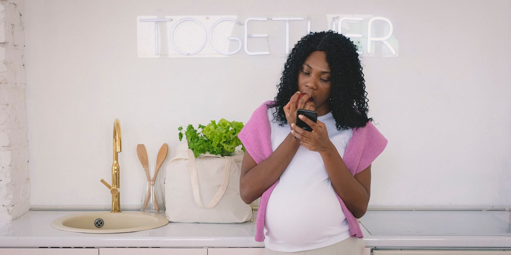 A pregnant woman uses her smartphone in a kitchen while eating an apple