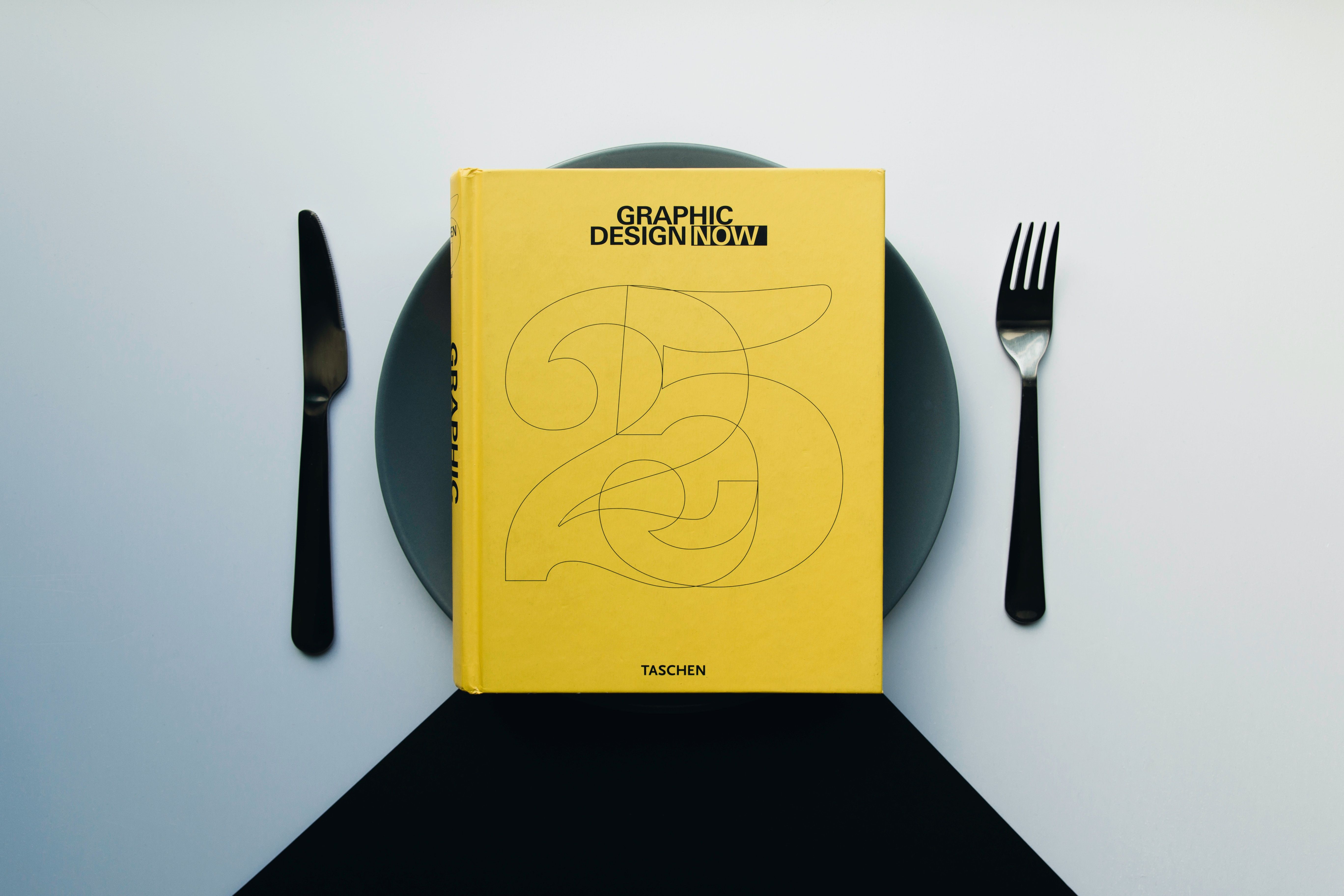 A simple, yellow graphic design book featured on a dinner plate with a knife and fork beside it.