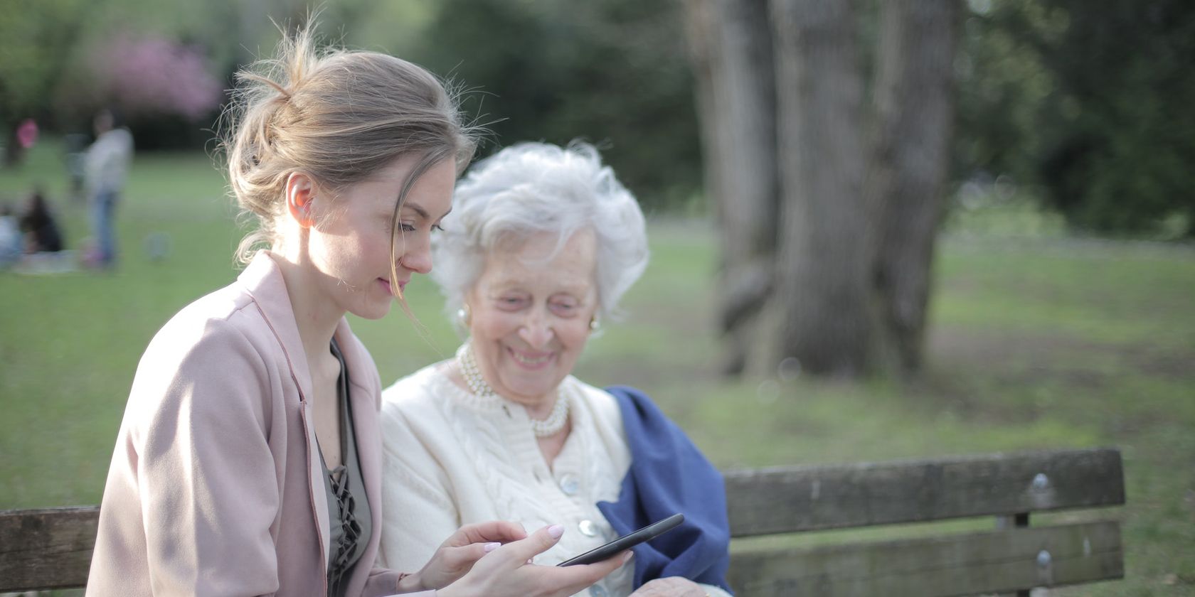 an elderly woman sitting on a park bench with a younger woman assisting them with a smartphone
