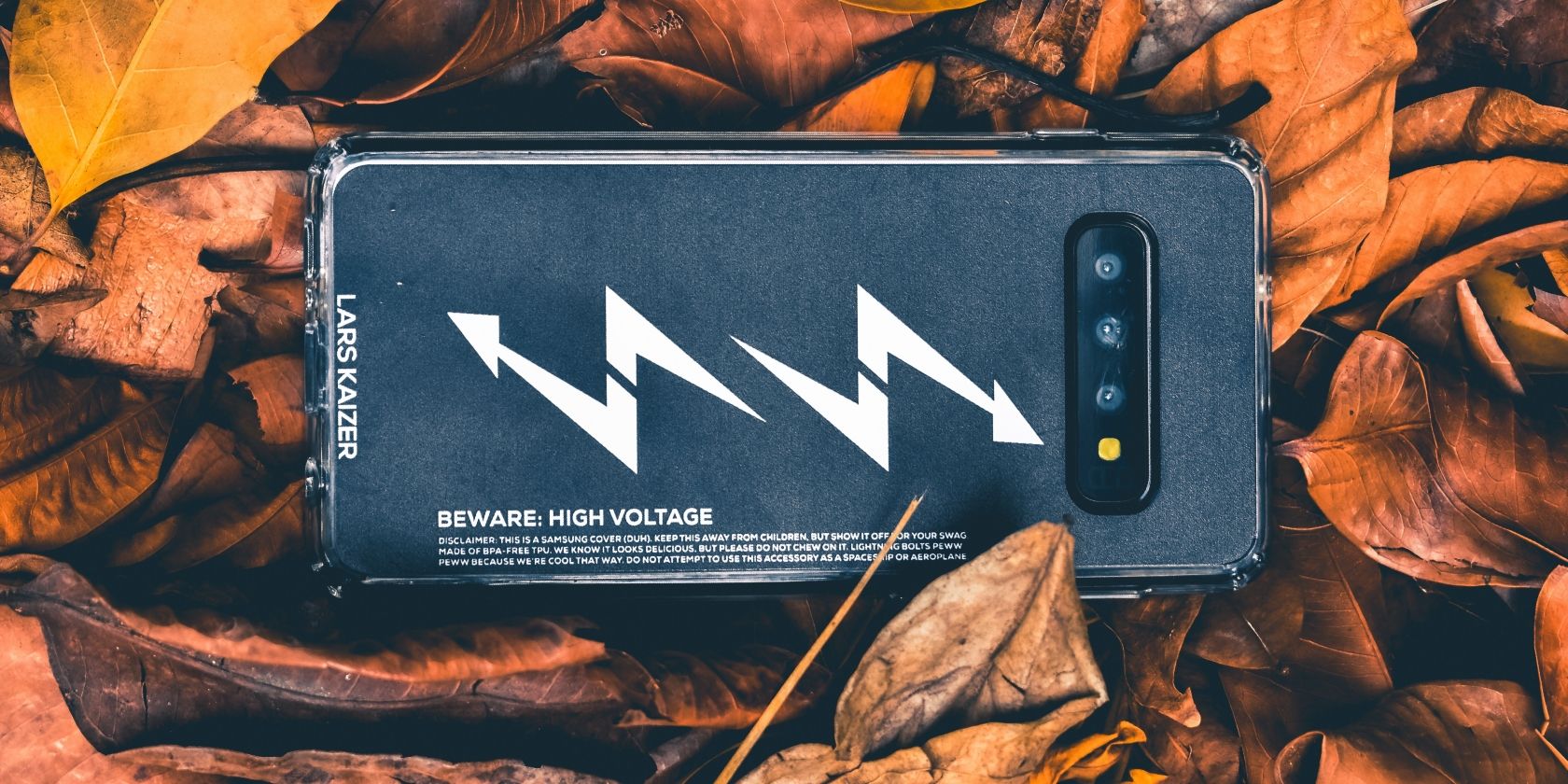 Samsung phone facedown on foliage, with a cover featuring a high voltage message on its back