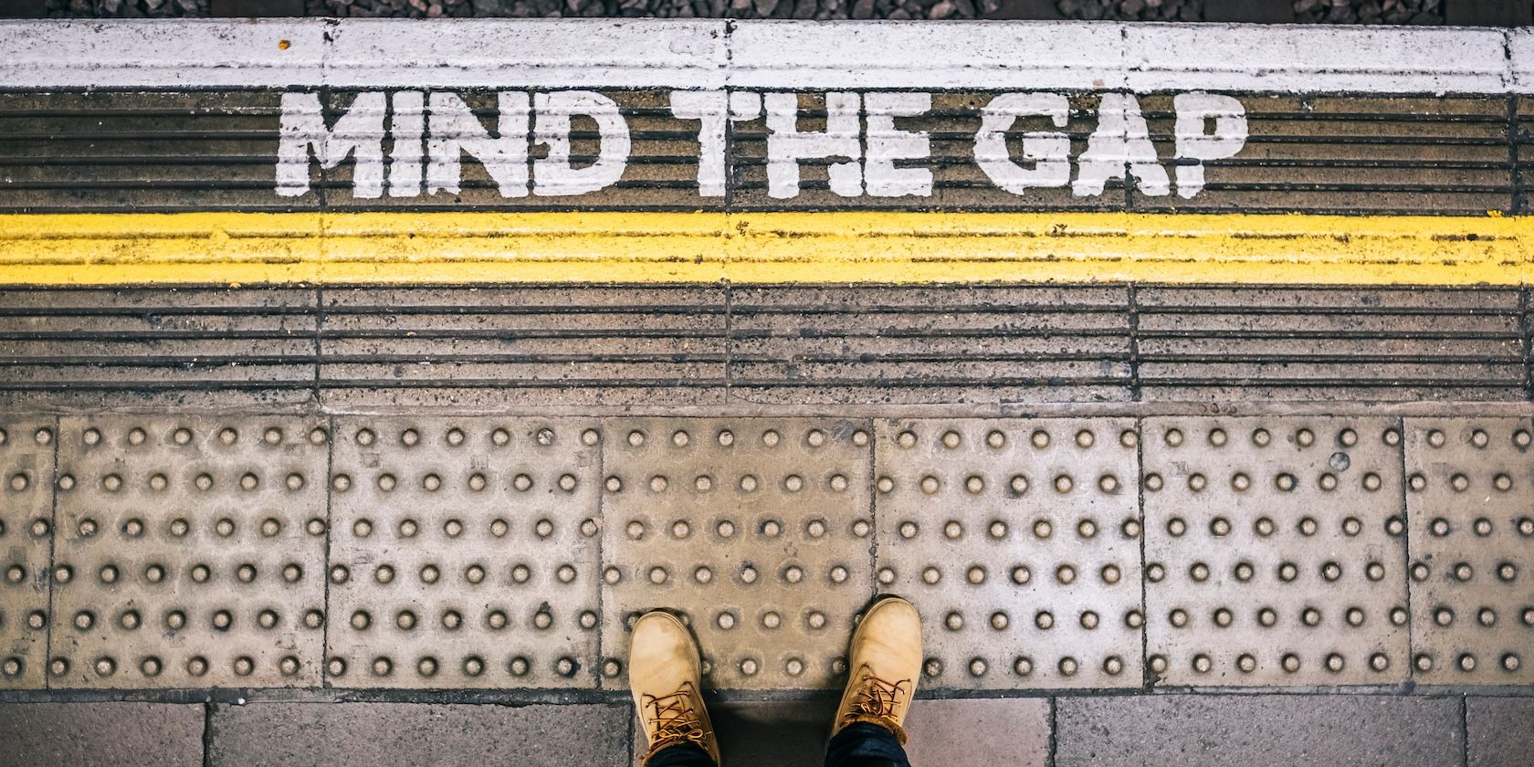 A pair of feet seen from above, near the edge of a station platform with “Mind The Gap” written on the floor
