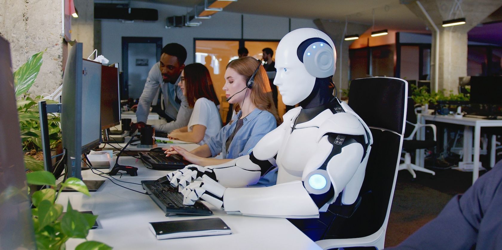 A robot sits at a desk appearing to type on a keyboard, working alongside human employees.