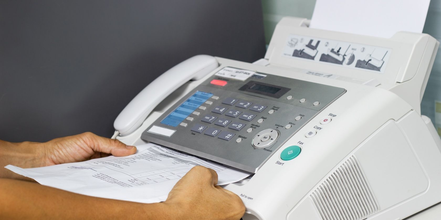 Hands feeding a piece of paper into a bulky fax machine with a telephone receiver and a panel of buttons.