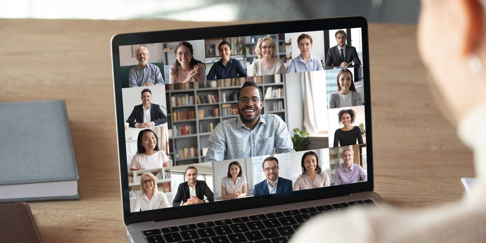 A laptop screen shows a video call with the portraits of many people in small thumbnails surrounding one person in close-up