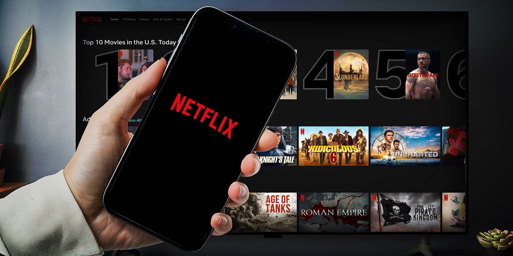A hand holding an iphone with the Netflix logo on the screen in front of a TV displaying the Netflix interface and top movies.
