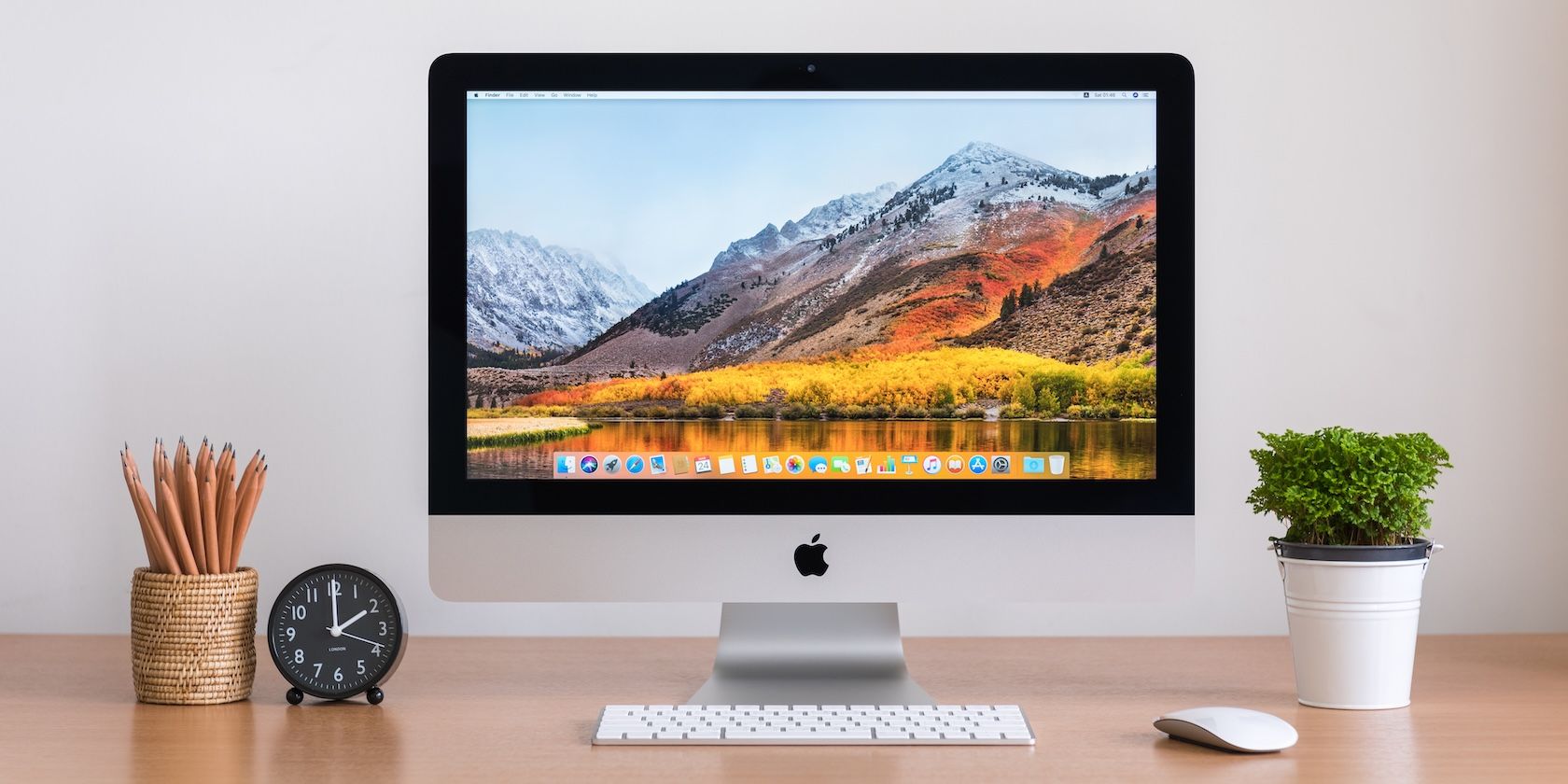 An iMac, showing the default macOS wallpaper, on a desk alongside a clock, pencil holder, plant, keyboard, and mouse.